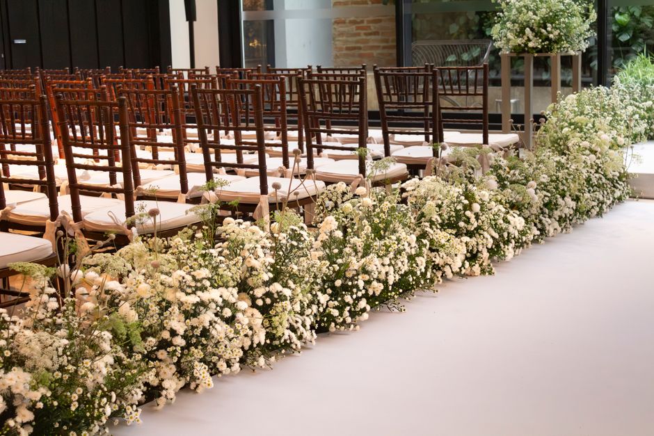 A floral arrangement featuring a dense border of white baby’s breath and small white roses, lining the aisle of an indoor wedding venue, with a row of wooden chairs with white cushions placed closel