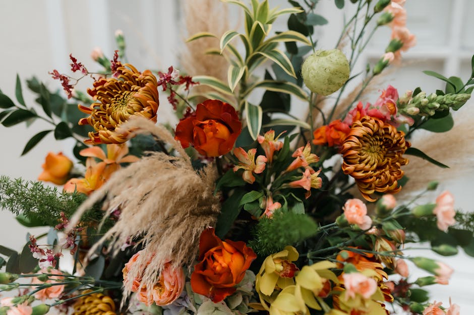 A vibrant floral arrangement featuring a mix of flowers including orange roses, large burnt-orange chrysanthemums, and delicate pink snapdragons, complemented by green foliage with variegated leaves a