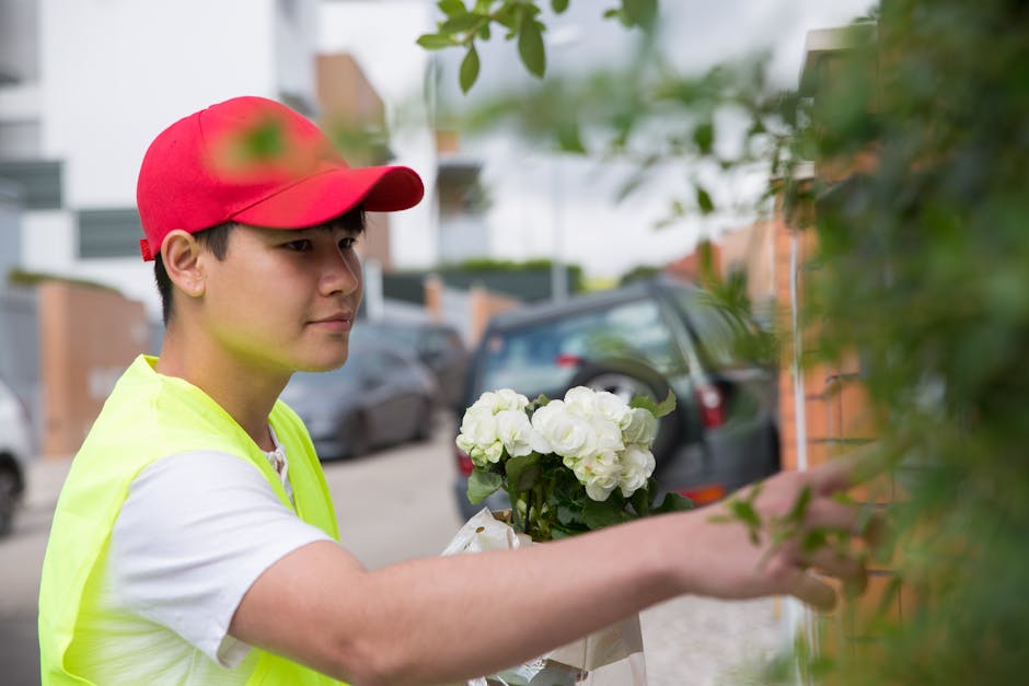 A young male flower delivery courier wearing a red cap and a high-visibility yellow vest is placing a bouquet of fresh white roses and green foliage against a brick wall. The bouquet is wrapped in cle