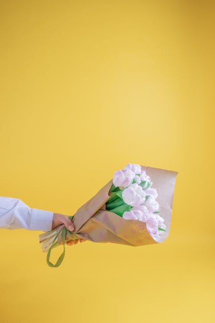 A person’s hand is visible, gently holding a bouquet of fresh, pastel pink and white tulips wrapped in light brown paper with a green ribbon. The tulips have smooth, rounded petals with some slightl