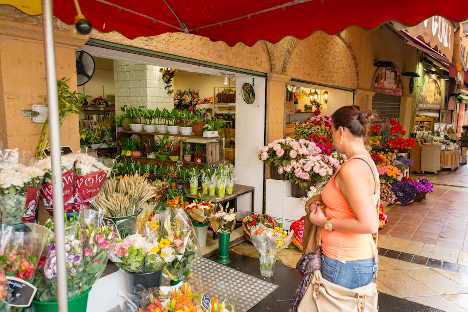 A woman stands in front of a flower shop under a red canopy, holding a small bouquet of flowers wrapped in brown paper. The shop's display includes a variety of fresh, vibrant flowers such as pink ros